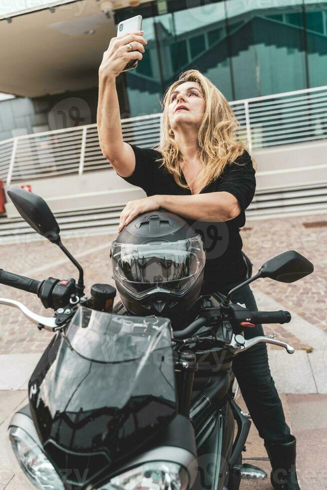 mitten åldrig blond kvinna cyklist är tar en selfie med smartphone ridning henne kraftfull svart motorcykel foto