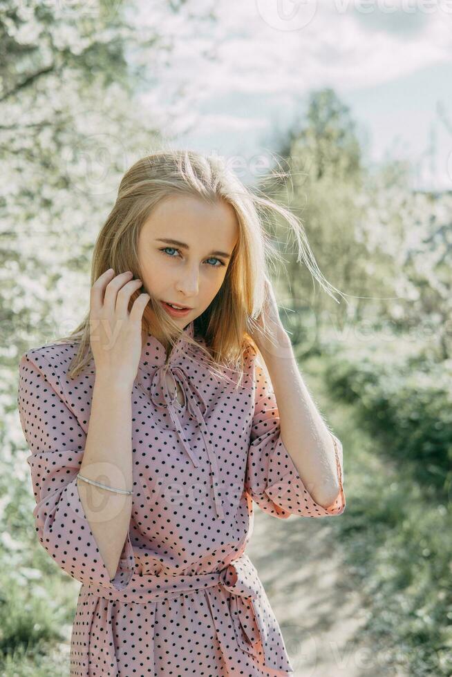 blond flicka på en vår promenad i de trädgård med körsbär blommar. kvinna porträtt, närbild. en flicka i en rosa polka punkt klänning. foto