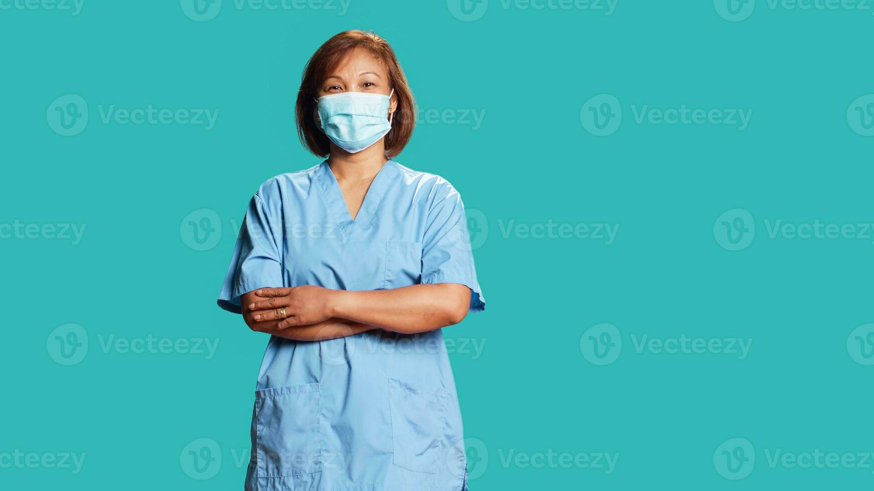 porträtt av glad asiatisk sjuksköterska stående med vapen vikta, isolerat över blå studio bakgrund. Lycklig bipoc kvinna bär skyddande klinisk ansikte mask till förhindra spridning av sjukdomar foto