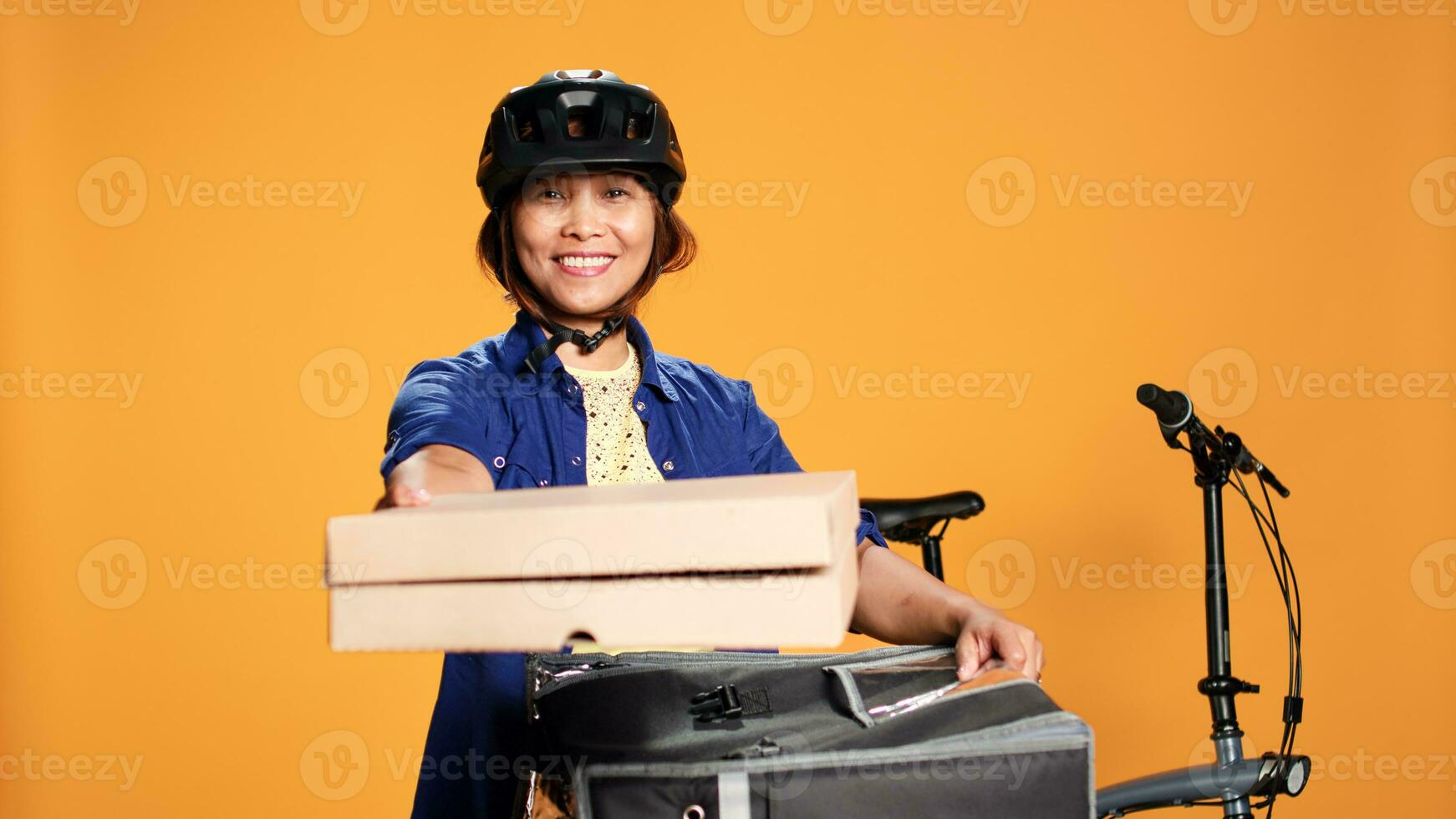 vänlig pizza leverans kvinna bär cykel redskap föra beställa till kund. optimistisk asiatisk kurir få lunch lådor ut av termisk mat ryggsäck, isolerat över orange studio bakgrund foto