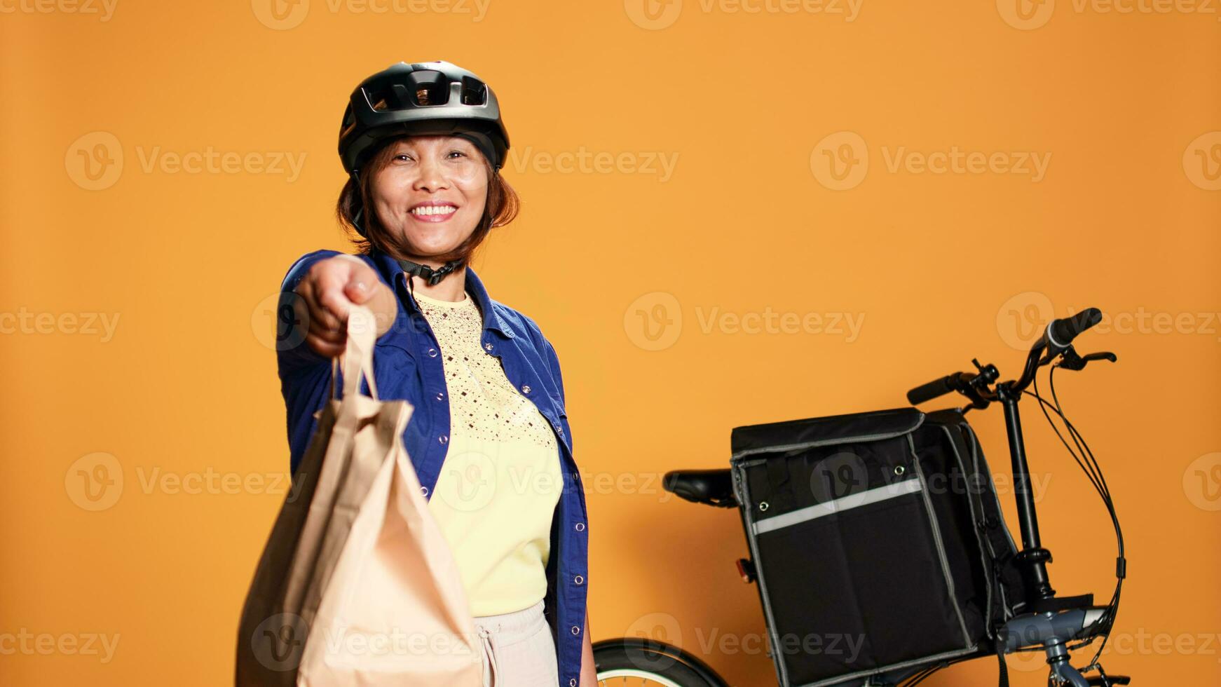 kurir väntar i främre av lägenhet dörr för kund till plocka upp hämtmat ordning. mat leverans cyklist erbjudande ta ut lunch väska till klient, isolerat över orange studio bakgrund foto