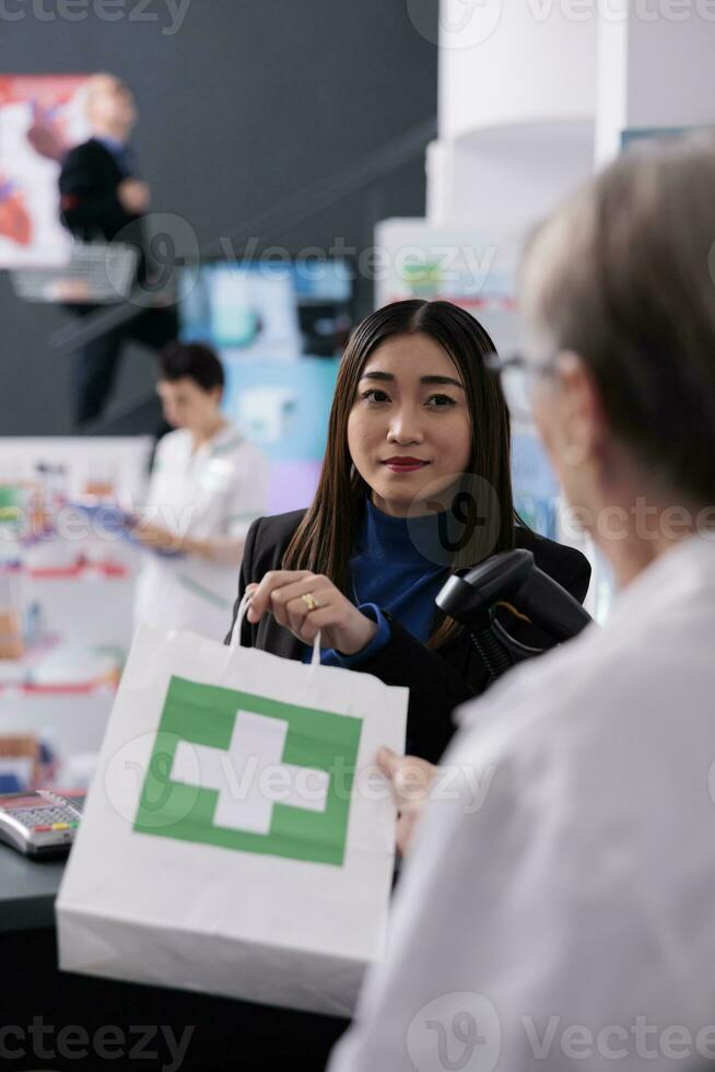 kvinna uppköp piller på apotek kontanter Registrera skrivbord och lyssnande till apotek kassör. farmaceutisk arbetstagare ger medicinsk Produkter handla väska till ung asiatisk kund på kolla upp foto