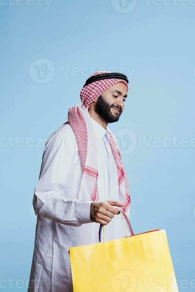 Lycklig konsument klädd i traditionell muslim kläder bärande gul handla väska. leende arab person bär thobe och ghutra slöja innehav Lagra inköp paket i studio foto