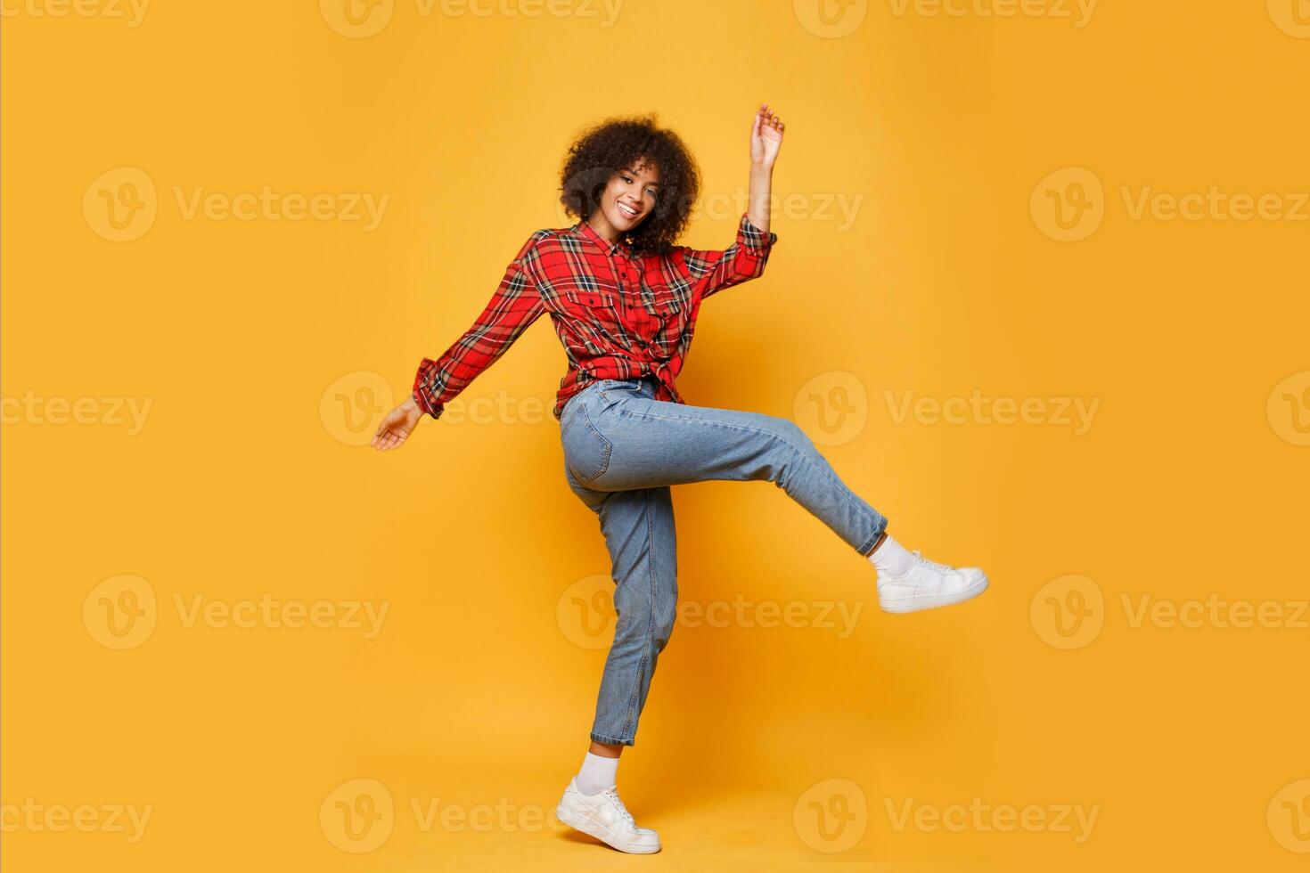 aktiva flicka med eleganta afro hårstrån dans på ljus orange bakgrund. studio Foto av Lycklig lockigt lady i röd skjorta Hoppar och har roligt inomhus. full längd.