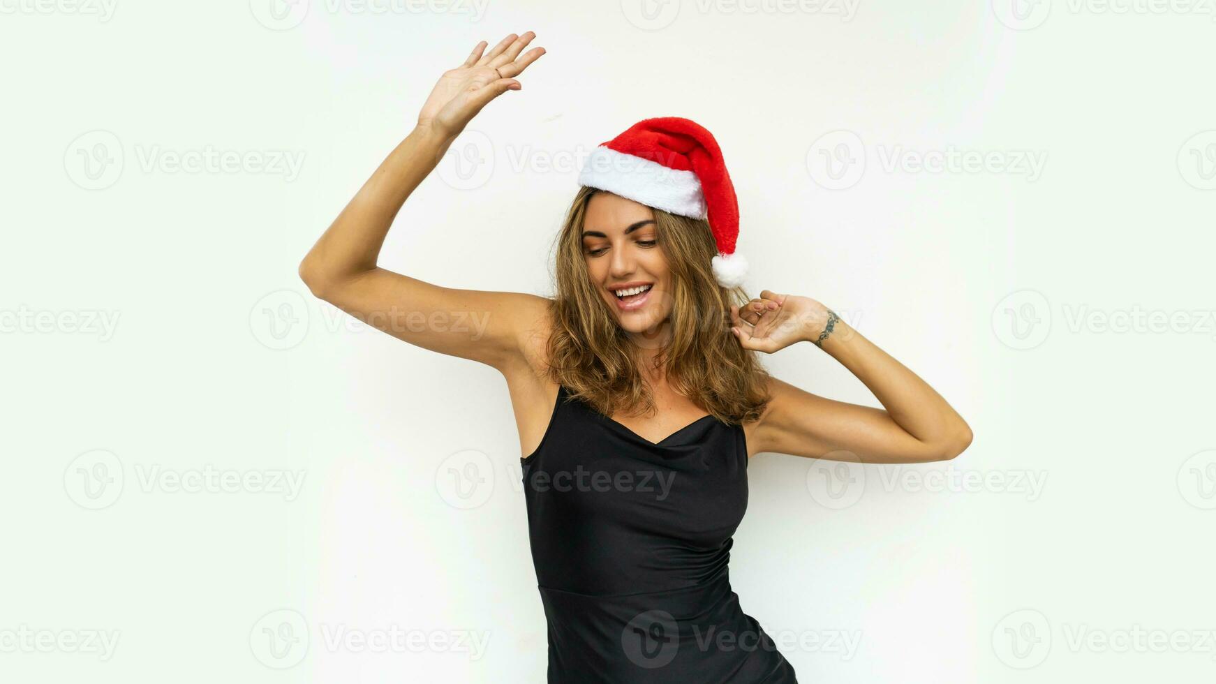sensuell kvinna i röd santa hatt och elegant svart klänning dans och har fest över vit bakgrund. foto