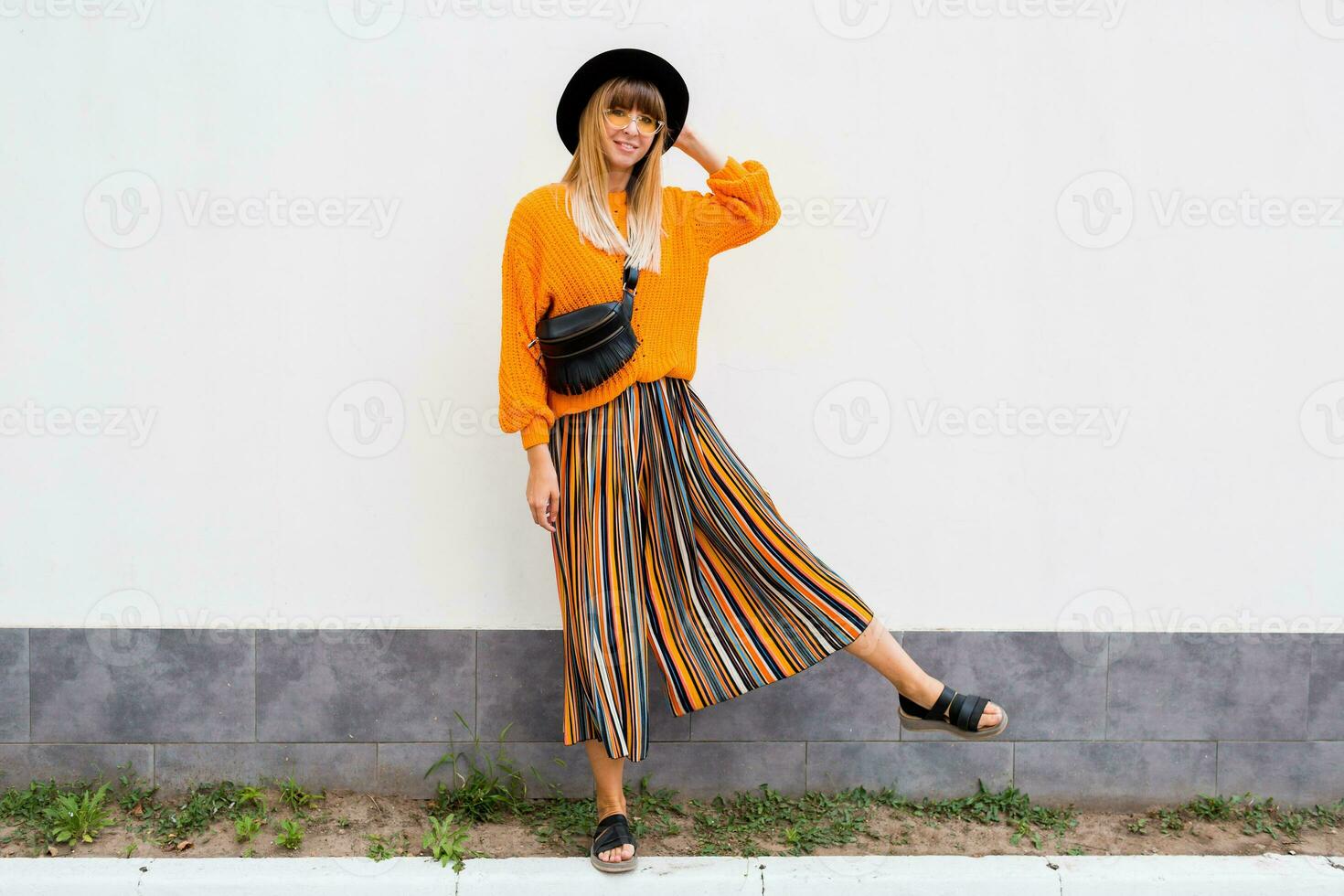 full längd bild av eleganta kvinna stående på vit bakgrund i eleganta orange Tröja och Flerfärgad rand culotte, oversize orange stickat Tröja, trendig luffare väska och hatt. foto