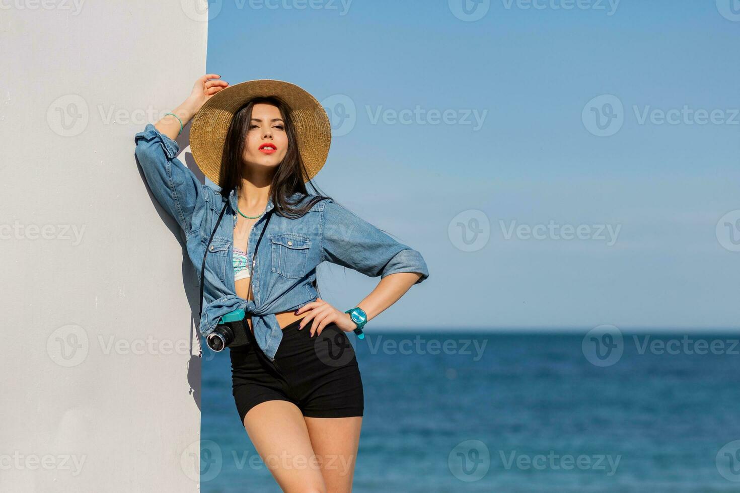 fantastisk kvinna med perfekt figur i sexig shorts och sugrör hatt Framställ på de strand. blå himmel och hav på bakgrund. foto