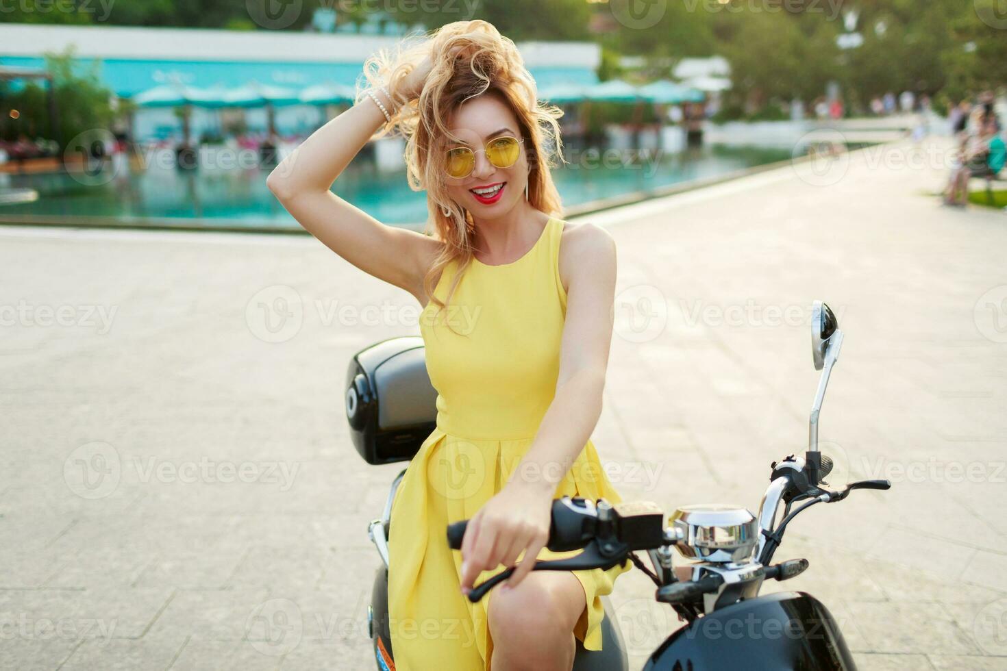 glad ingefära kvinna i gul glasögon och årgång klänning Sammanträde på eleganta motorcykel. semester humör. solig sommar dag. röd mun. foto