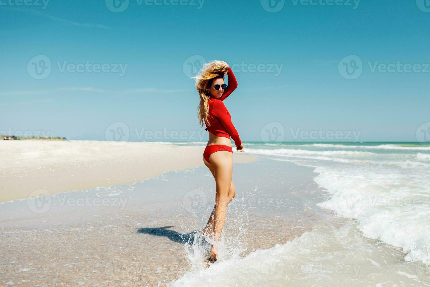 strand semester. varm skön kvinna i röd bikini stående med henne vapen Uppfostrad till henne huvud njuter ser se av strand hav på varm sommar dag. foto