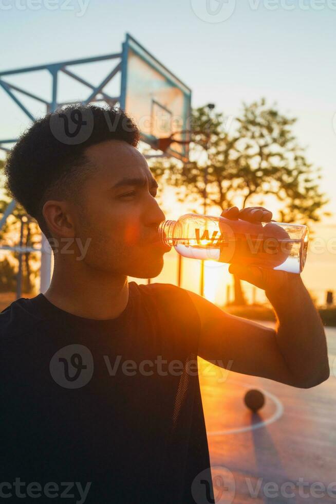 svart man håller på med sporter i morgon, dricka vatten på basketboll domstol på soluppgång foto