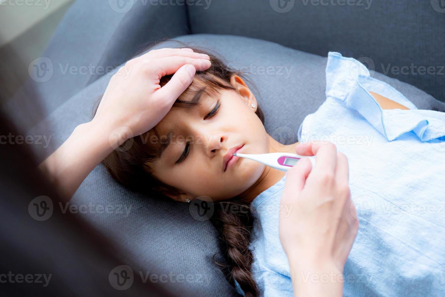 mamma hand tar temperaturen med flickan är en sjuk som ligger på soffan foto