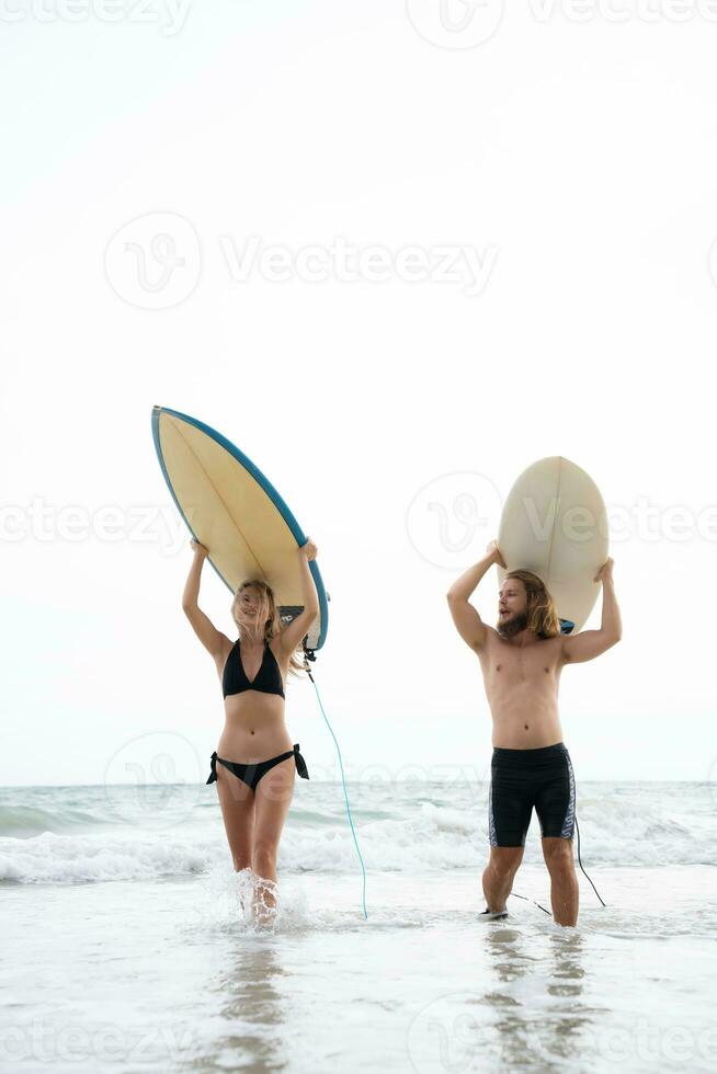 ung man och kvinna innehav surfingbrädor på deras huvuden och promenad in i de hav till surfa foto