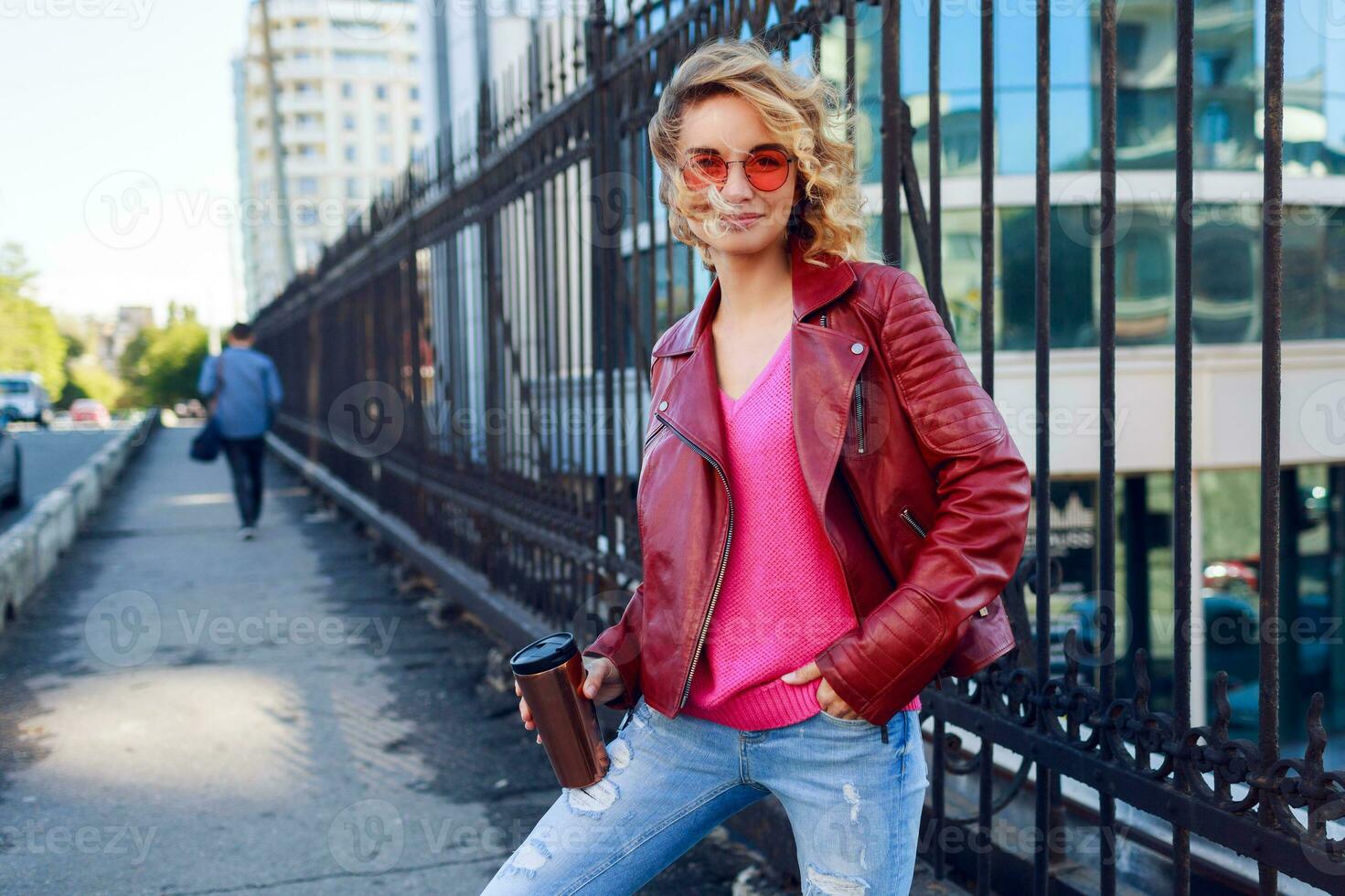 Lycklig blond kvinna Framställ på modern gator, dricka kaffe eller cappuccino. eleganta höst utrusta, läder jacka och stickat Tröja. urban bakgrund. rosa solglasögon. foto