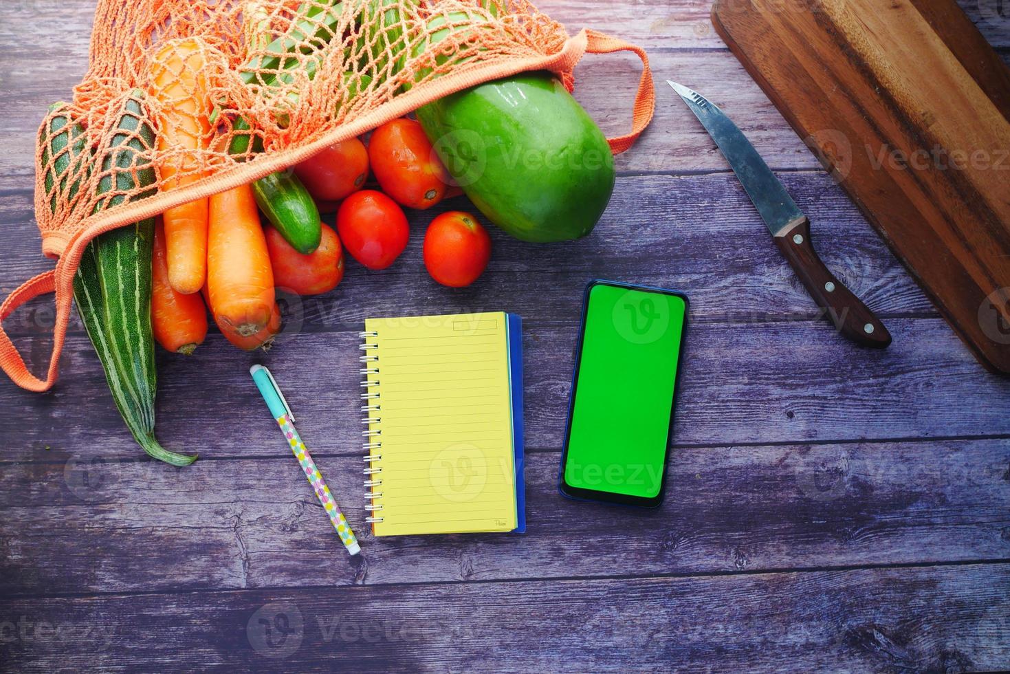 färska grönsaker, skärbräda, anteckningsblock och smart telefon på bordet foto