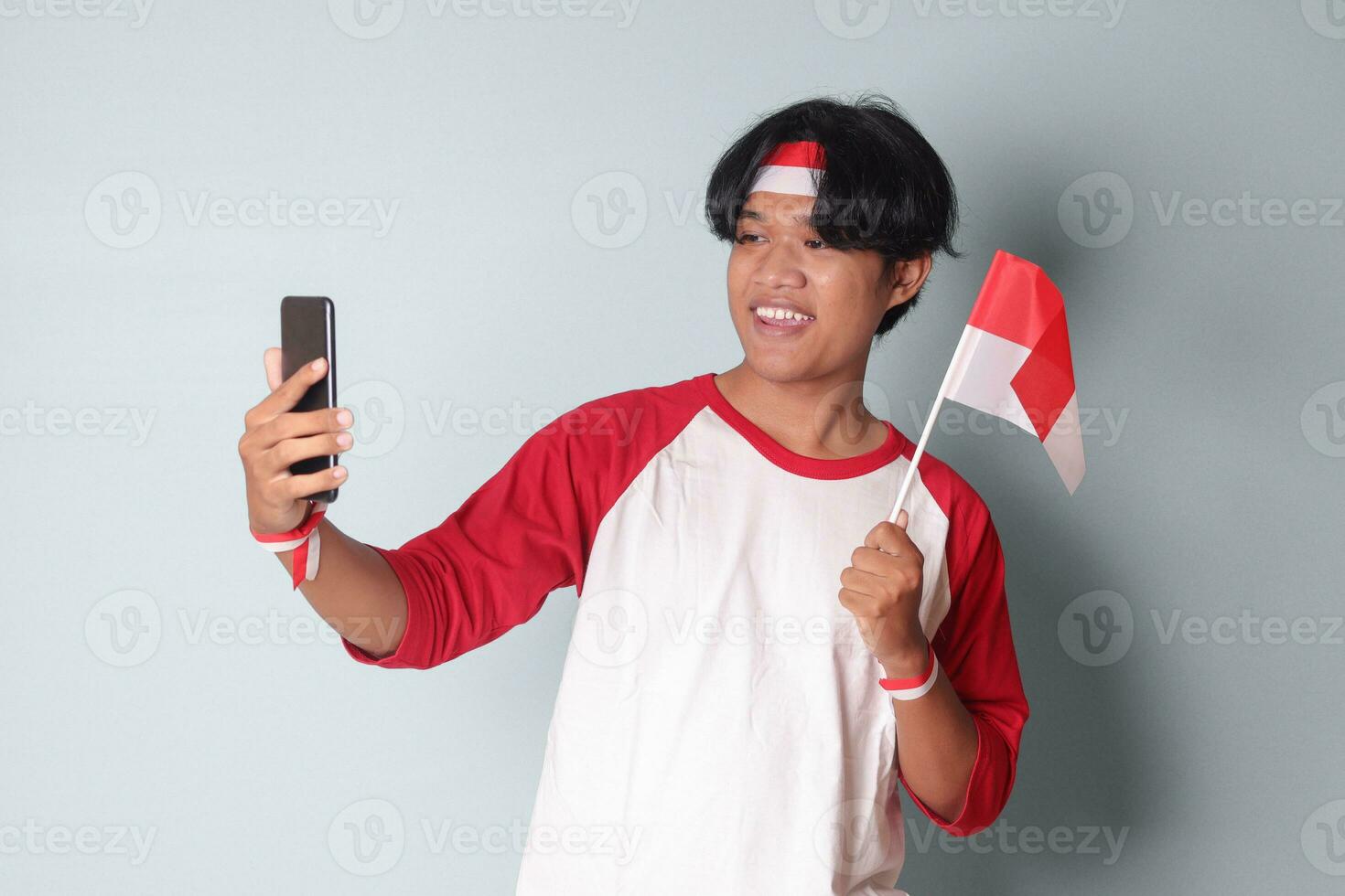 porträtt av attraktiv asiatisk man i t-shirt med röd och vit band på huvud, tar en bild av han själv medan innehav indonesien flagga. isolerat bild på grå bakgrund foto