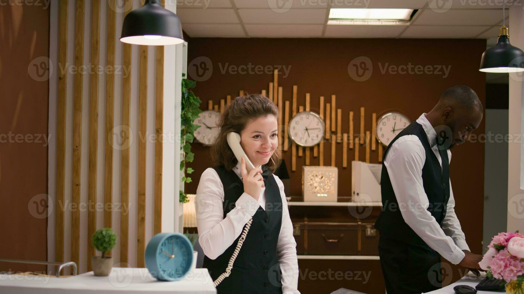 hotell personal svar samtal på reception, använder sig av fast telefon telefon till göra avlägsen bokningar på främre skrivbord. vänlig receptionist förvaring bevis av boende och rum reservationer. foto