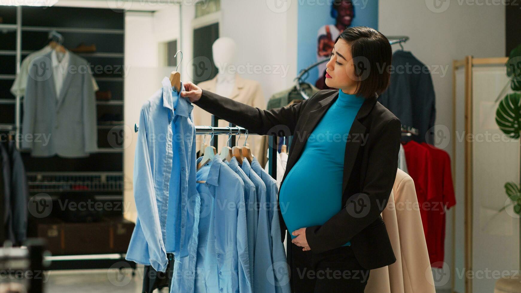 asiatisk kvinna med bebis stöta uppköp trendig shirts i mode boutique, gravid kund ser på kläder på galgar. kvinna klient håller på med handla i Kläder Lagra. stativ skott. foto