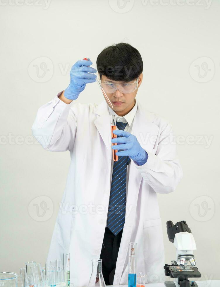 porträtt asiatisk man studerande forskare bär en läkare klänning i de labb ser hand på kemist. orsakade förbi blandning reagens i vetenskaplig forskning laboratorier med testa rör och mikroskop på de tabell foto