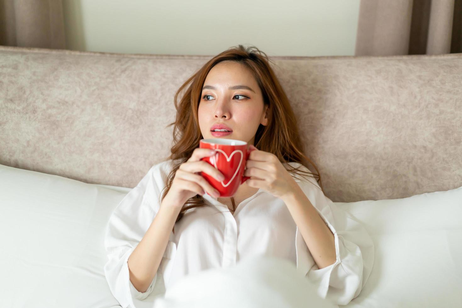 porträtt vacker asiatisk kvinna vaknar och håller kaffekopp eller mugg på sängen på morgonen foto