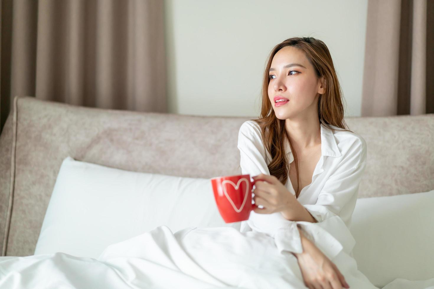 porträtt vacker asiatisk kvinna vaknar och håller kaffekopp eller mugg på sängen på morgonen foto