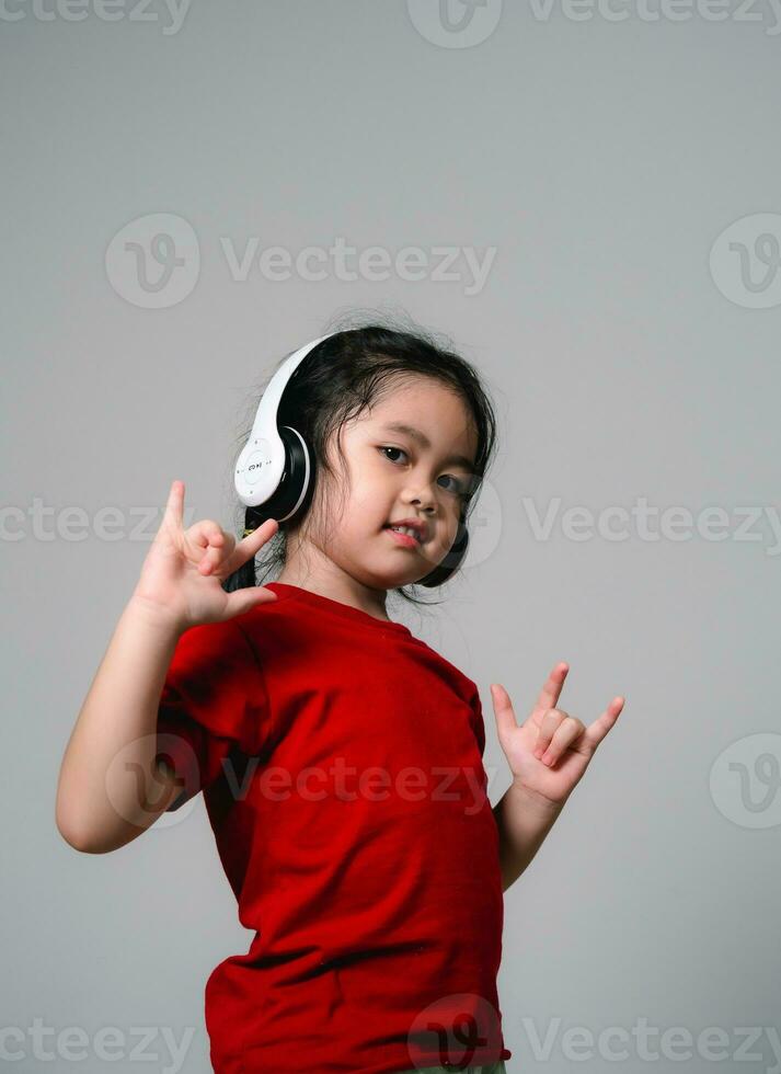 glad Lycklig asiatisk barn flicka med svart hår lyssnande till musik låtar i hörlurar på en grå bakgrund. porträtt söt flicka krigande röd skjorta och ser på kamera studio belysning. foto