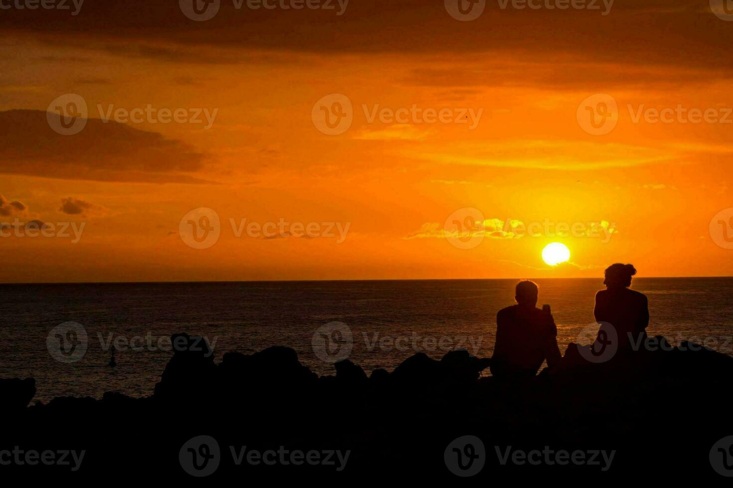 två människor Sammanträde på stenar tittar på de solnedgång foto