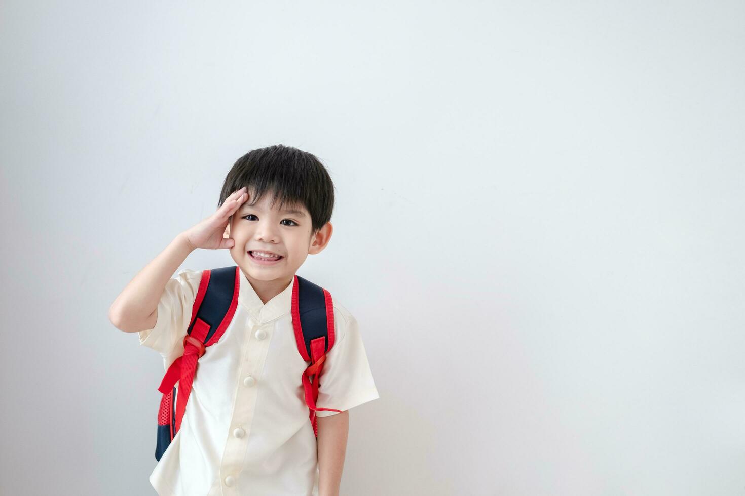 asiatisk pojke bär traditionell thai Kläder, stående med en skola väska. låtsas till säga Hej och få redo för skola på en vit bakgrund foto