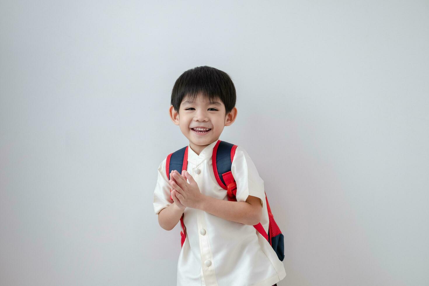 asiatisk pojke bär traditionell thai Kläder, stående med en skola väska. låtsas till säga Hej och få redo för skola på en vit bakgrund foto