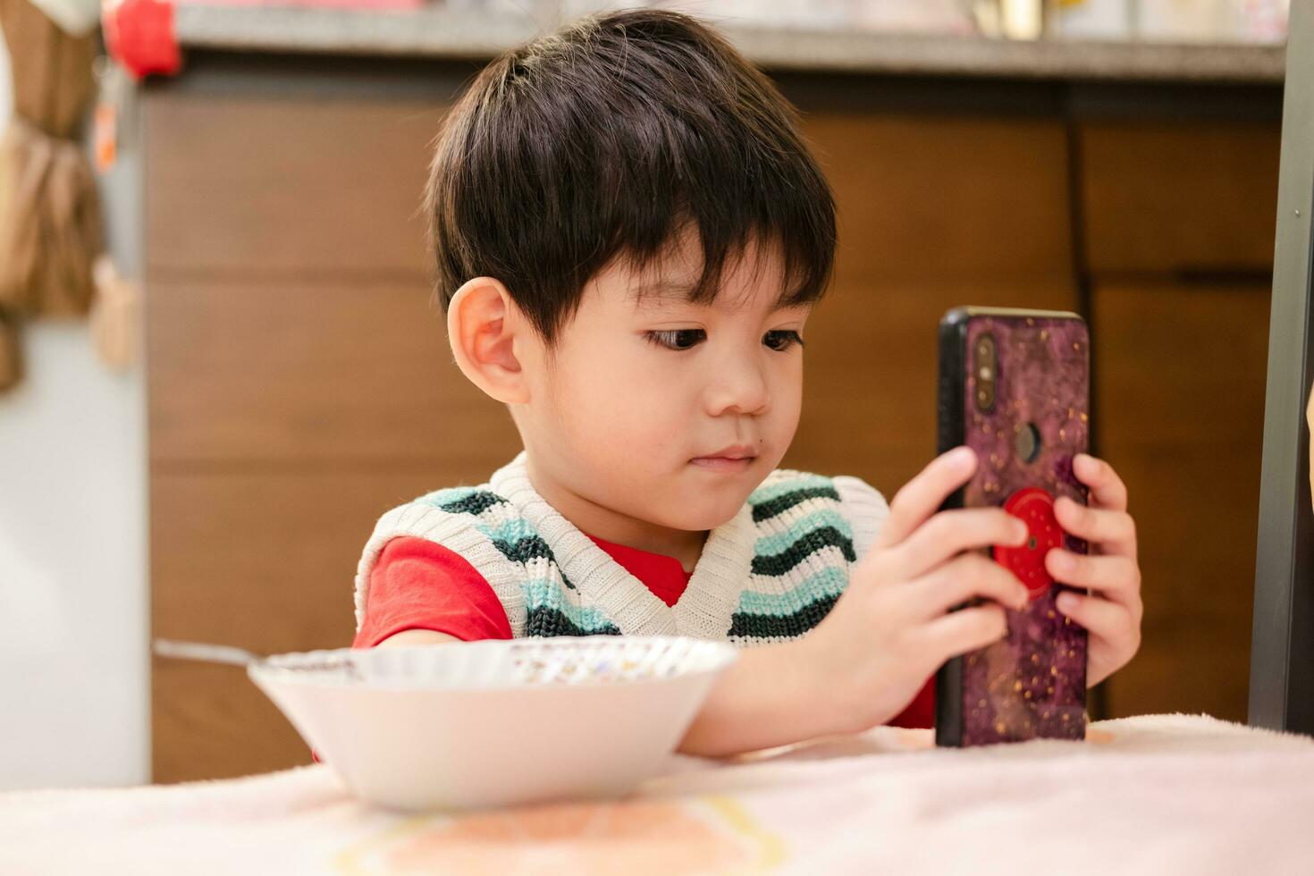 de ansikte av ett asiatisk barn äter medan ser på de telefon foto