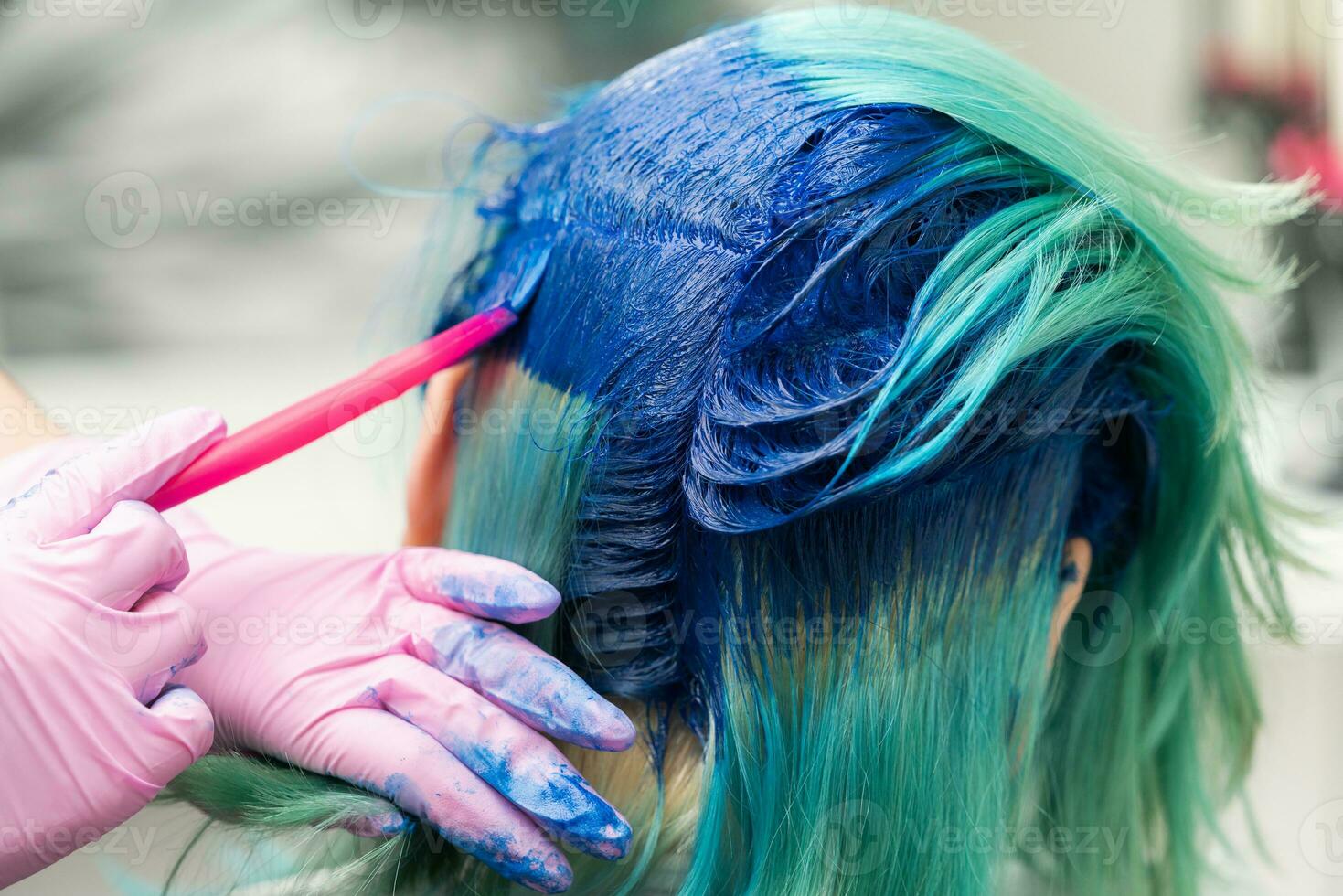 sida se av bearbeta av färgning hår i unik Färg. frisör i handske använder sig av rosa borsta medan applicering blå måla till kund med smaragd- hår Färg foto