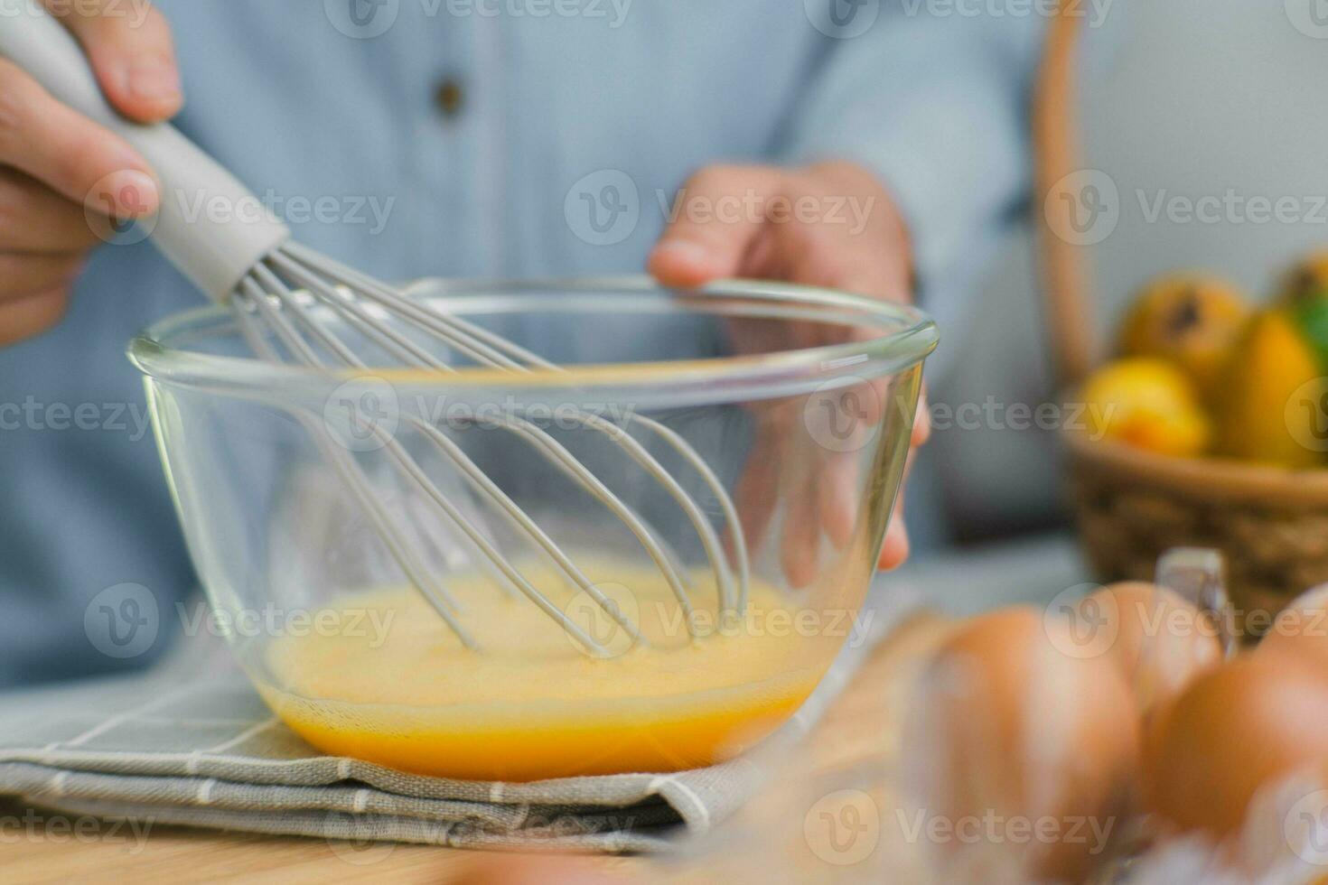 ung kvinna matlagning i ljus kök, händer vispning ägg i en skål placerad på handduk och trä- tabell. framställning Ingredienser för friska matlagning. hemlagad mat foto