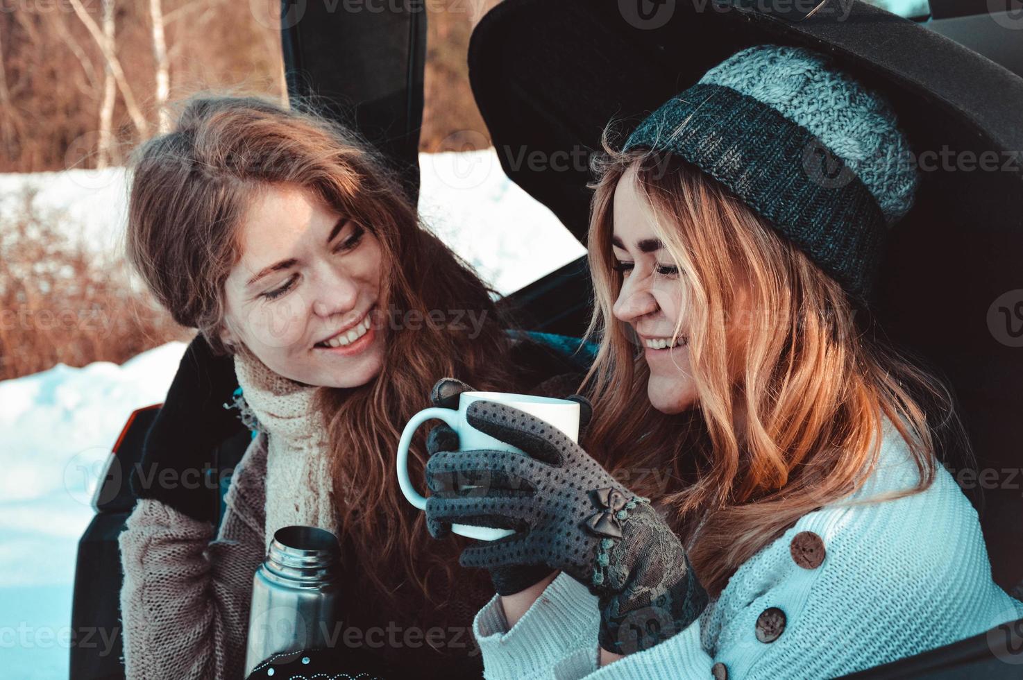 glada vänner i vinterskogen. två tjejer sitter i bagaget och dricker kaffe foto