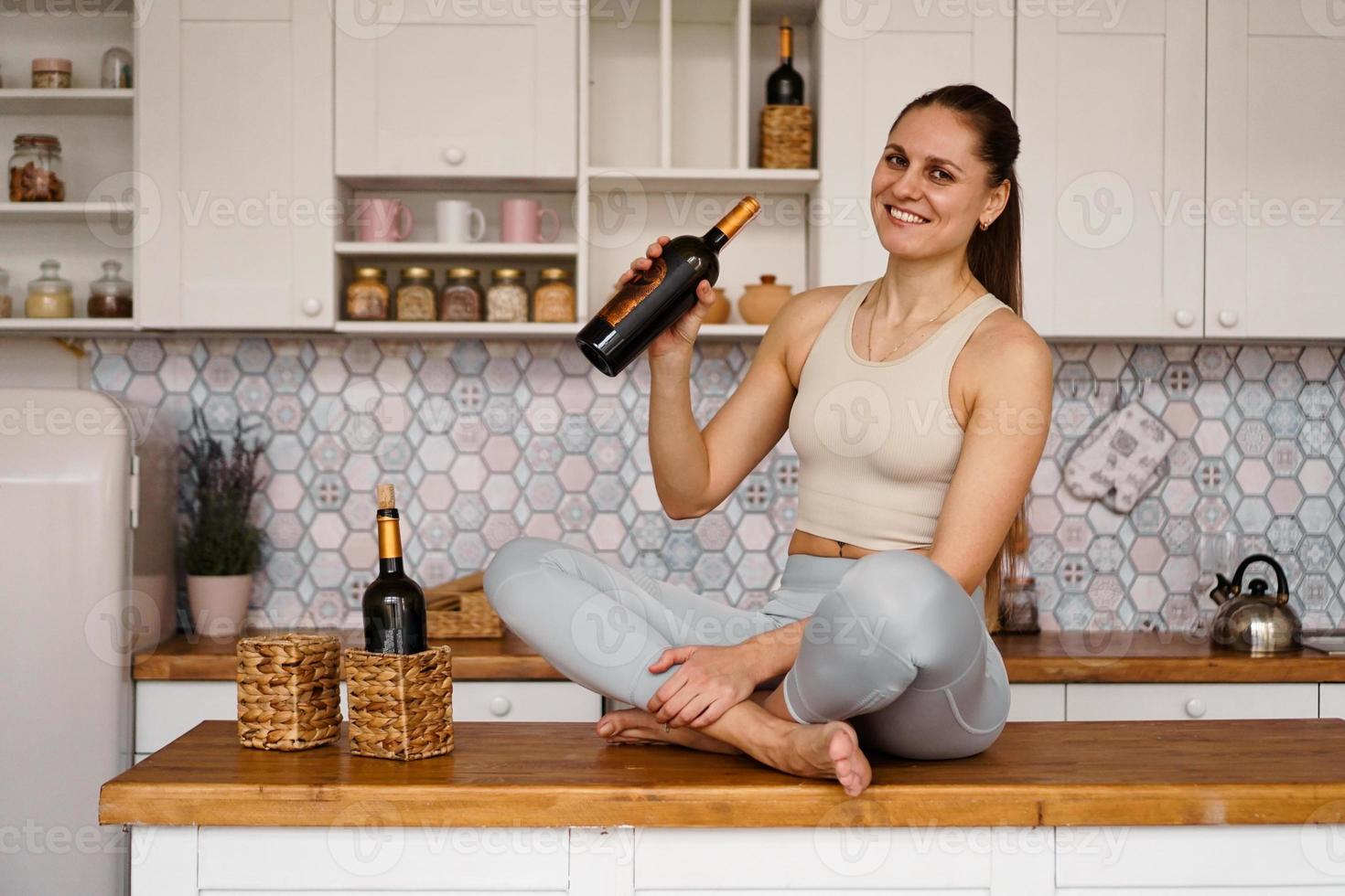 atletisk kvinna i en träningsdräkt i ett ljust kök dricker rött vin foto