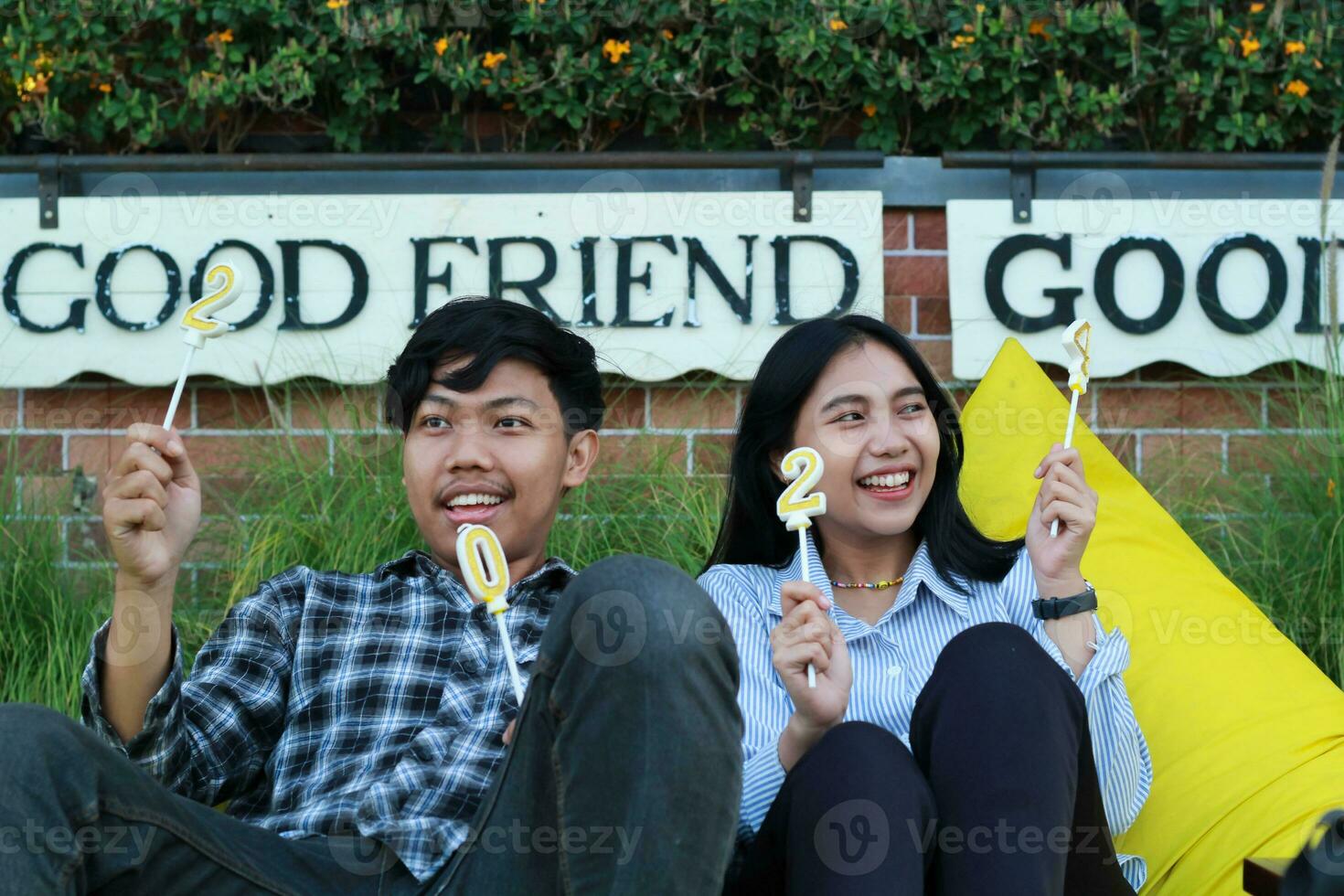 Lycklig asiatisk par tonåring höjning siffra 20 24 gyllene ljus medan Sammanträde på de gul böna väska i tillbaka gård med tegel vägg bakgrund, se bort foto