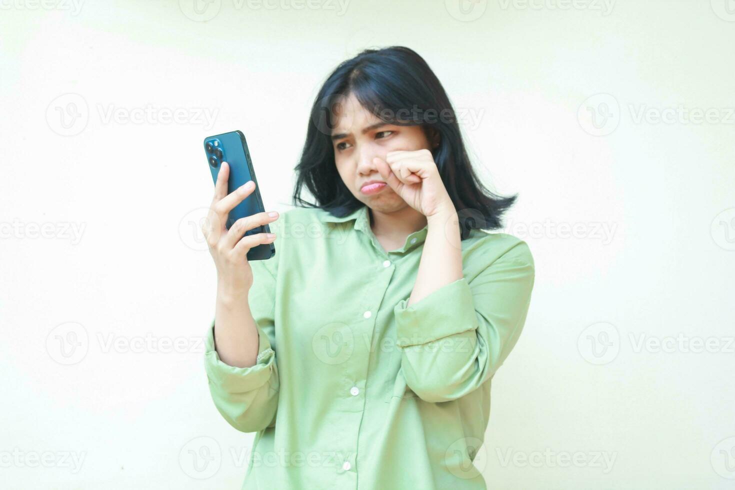 ledsen asiatisk kvinna våtservetter henne tårar efter gråta för använder sig av social media på smartphone håll på händer bär grön överdimensionerad skjorta stående över isolerat bakgrund. mental hälsa begrepp, mobbning foto