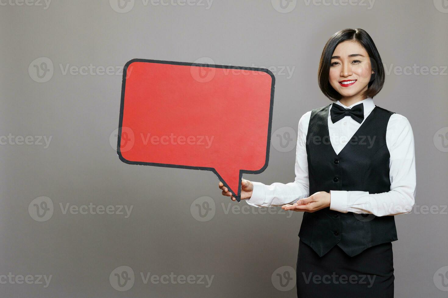 asiatisk servitris bär professionell enhetlig som visar röd tom tala ram studio porträtt. restaurang leende kvinna arbetstagare innehav tömma chatt bubbla attrapp och ser på kamera foto