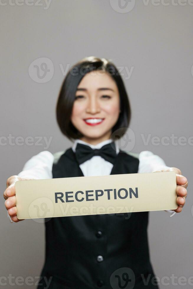 glad asiatisk restaurang servitris visa upp rostfri stål reception tallrik porträtt. leende hotell administratör som visar receptionist namn Skyltning och ser på kamera foto