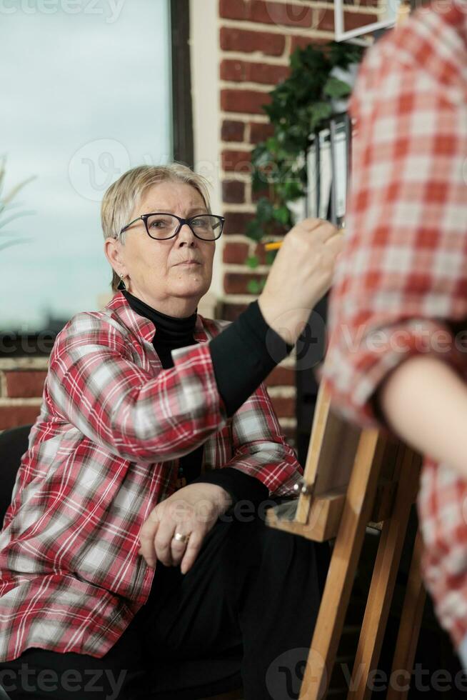 mogna kvinna inlärning till dra, pensionerad lady innehav penna, teckning på staffli och lyssnande till lärare, utvecklande konstnärlig Kompetens i pensionering. kreativ hobbies och konst terapi för seniors foto