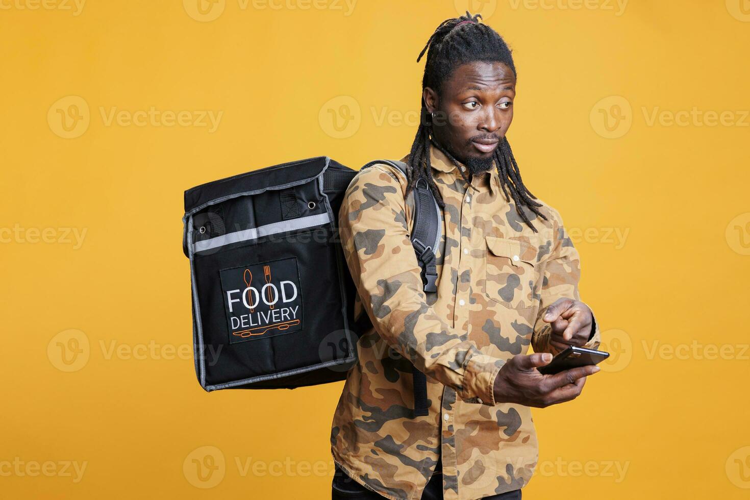 deliveryman kontroll vägbeskrivning och beställa detaljer på smartphone, bärande termisk ryggsäck i studio över gul bakgrund. afrikansk amerikan kurir leverans ta ut lunch. mat service foto