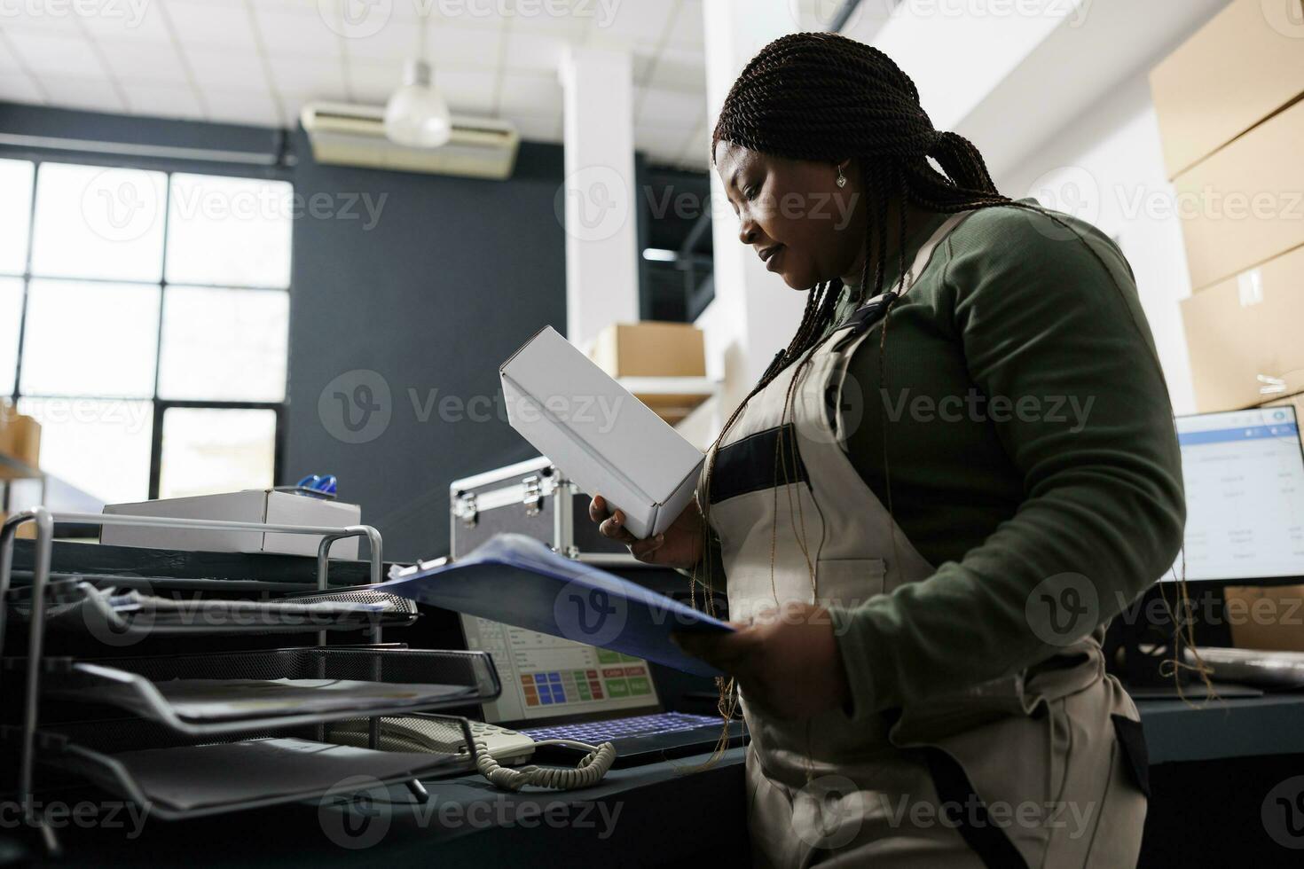 afrikansk amerikan arbetstagare ser på vit kartong låda, framställning kunder order på disken skrivbord i magasin. lager anställd bär industriell övergripande under varor lager foto