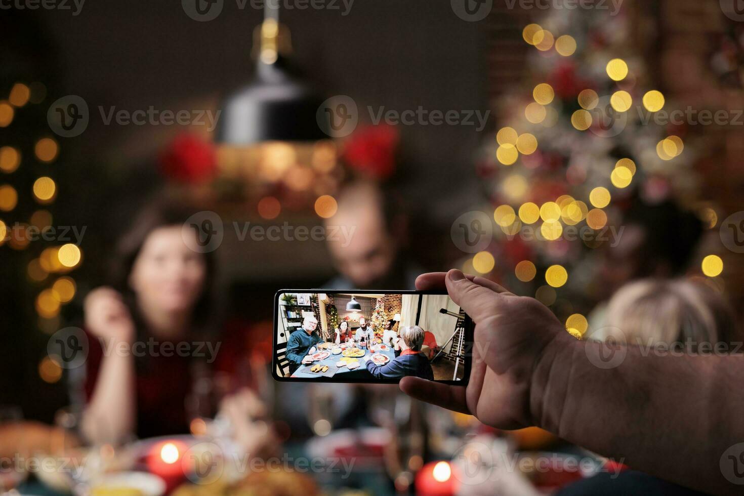 person tar foton av människor på tabell njuter middag fest med glasögon av alkohol omgiven förbi xmas träd och ornament. olika vänner och familj Framställ för bilder på jul eve.