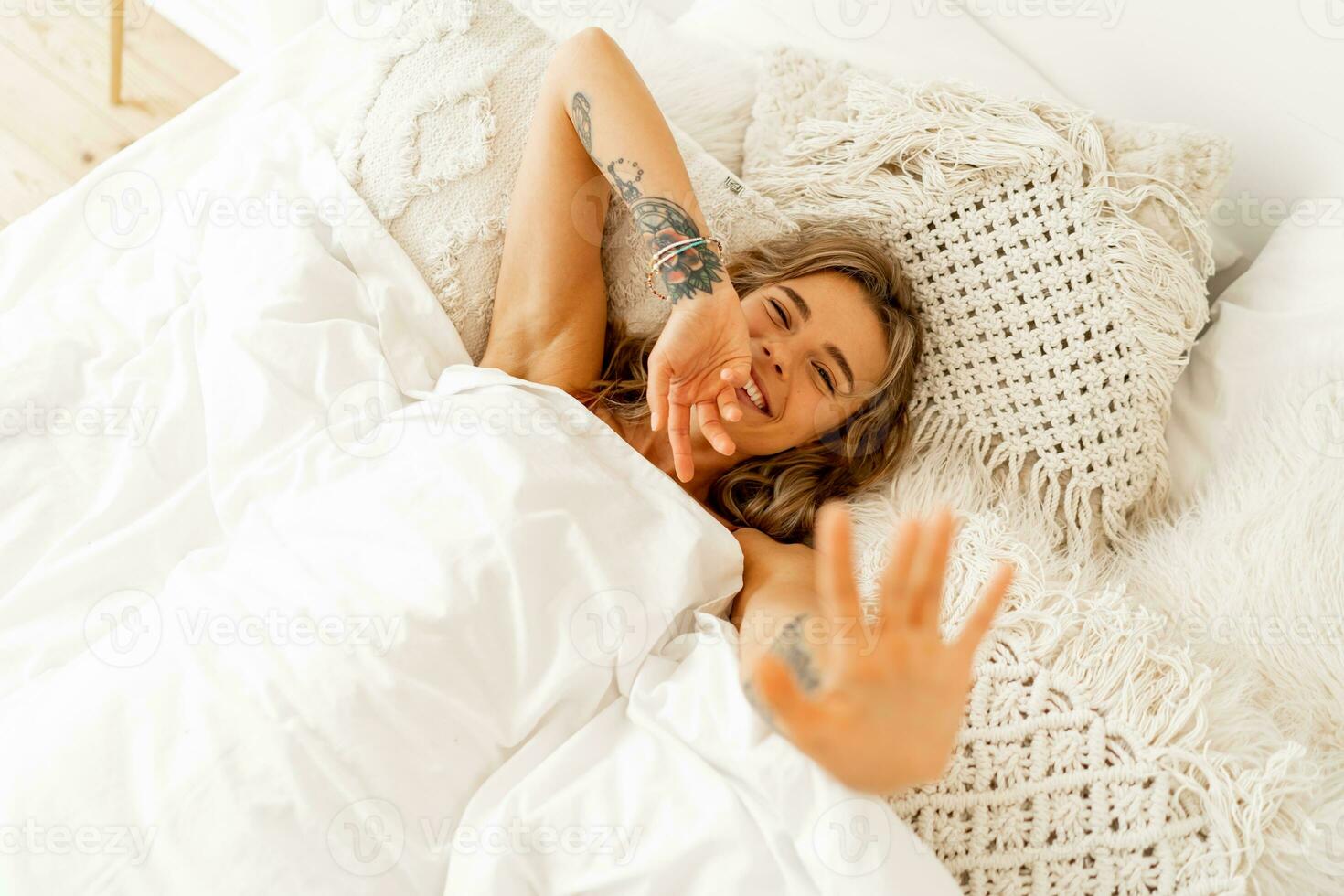 topp se porträtt av Lycklig glad kvinna liggande i vit mysigt säng vakna upp i de morgon, drar händer fram. värma färger, boho interiör. foto