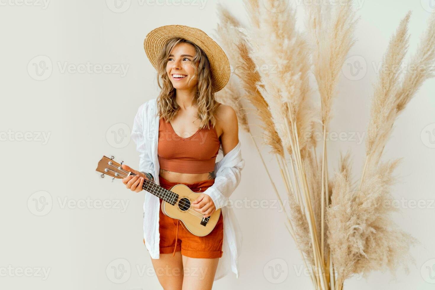 inomhus- Foto av blond graciös kvinna i sommar utrusta med sugrör hatt spelar ukulele guitare över vit bakgrund i studio med pampas gräs dekor.