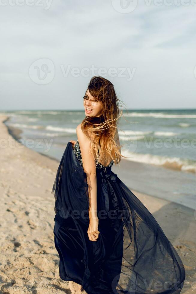 spännande graciös kvinna i elegant lång blå klänning lurar runt om och har roligt på de tropisk strand. semester tid foto