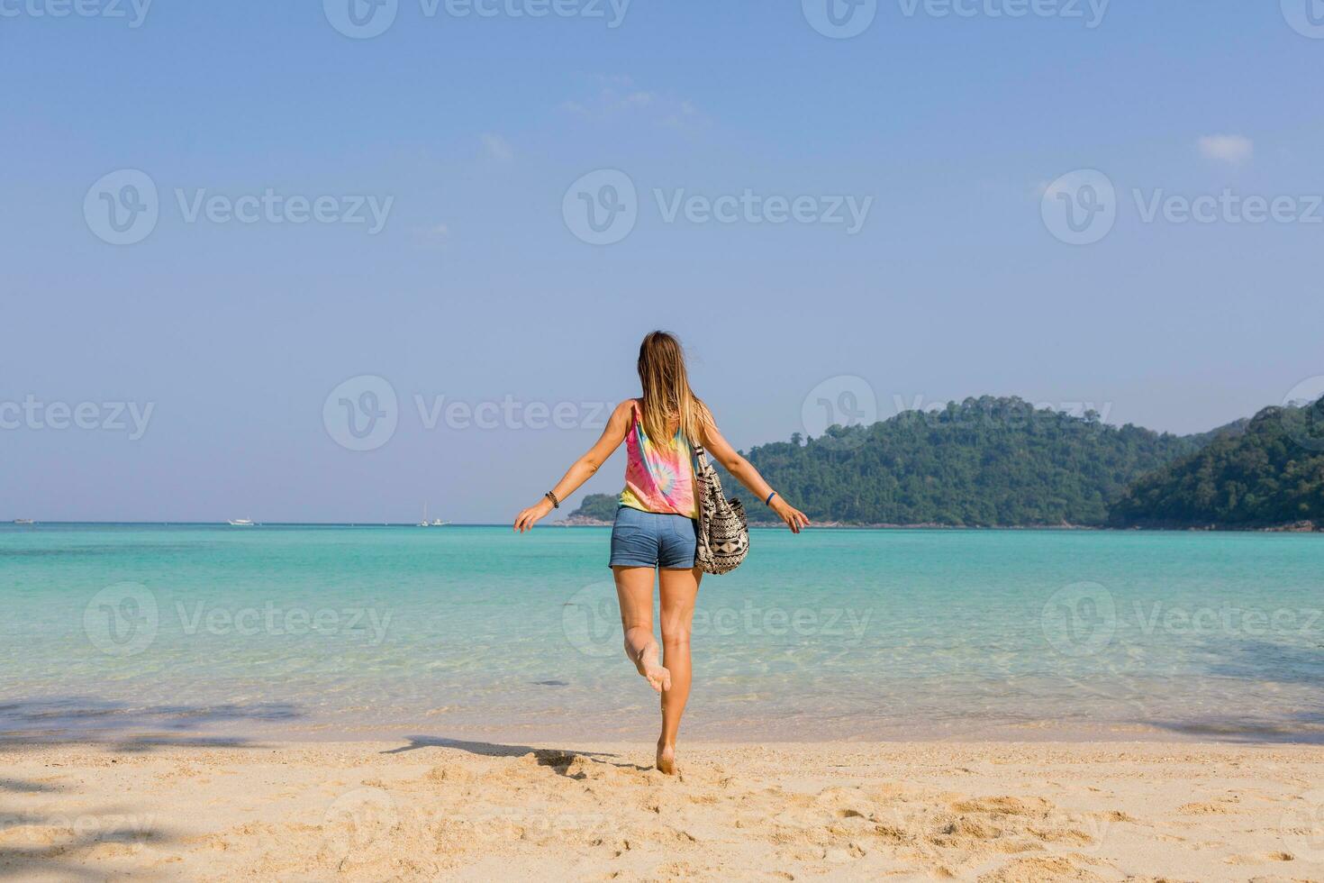 full längd porträtt från tillbaka av solbränd ung kvinna ser på Fantastisk blå hav och bergen se , njuter högtider. bär strand väska och färgrik utrusta. foto