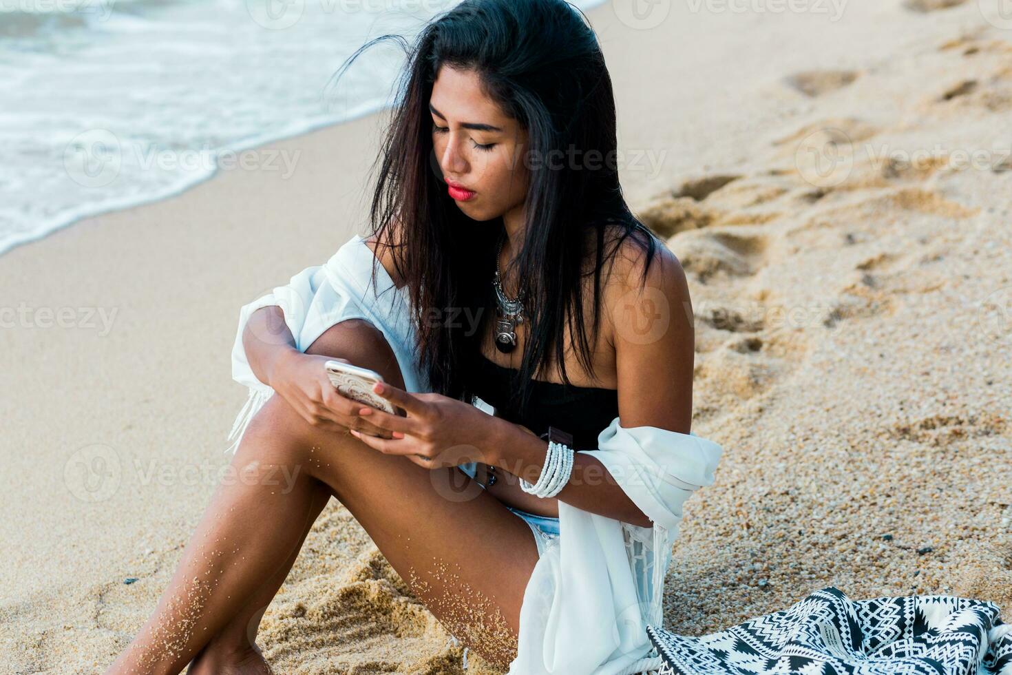 ung Söt Asien kvinna innehav och använder sig av mobil telefon testning på tropisk strand. bär eleganta vit boho omslag upp och trendig Tillbehör . röd mun. foto