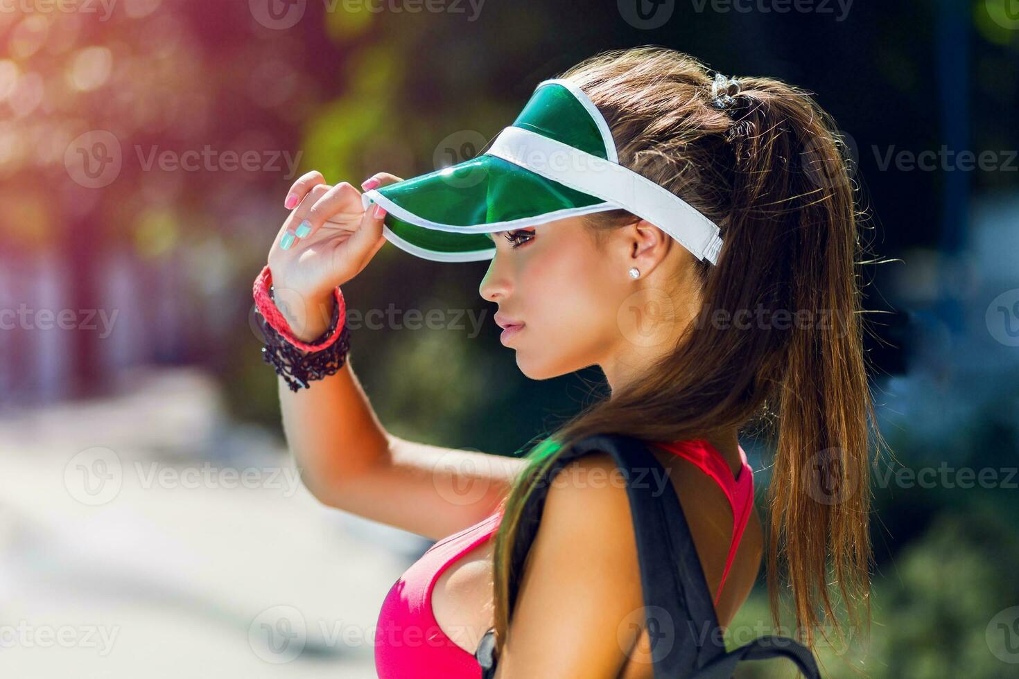 mode porträtt av ung skön kvinna i eleganta sporter enhetlig med neon ryggsäck och transparent visir går till spela tennis i värma solig sommar dag. foto