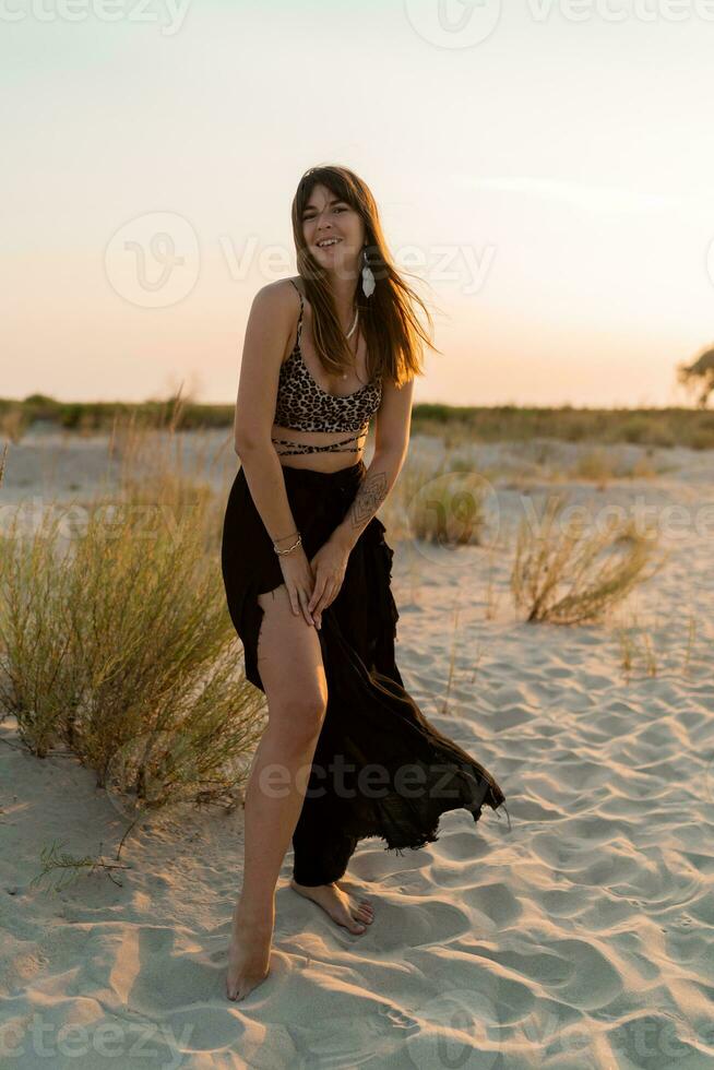 glad brunett kvinna i eleganta sommar boho utrusta Framställ på de strand. solnedgång färger. foto