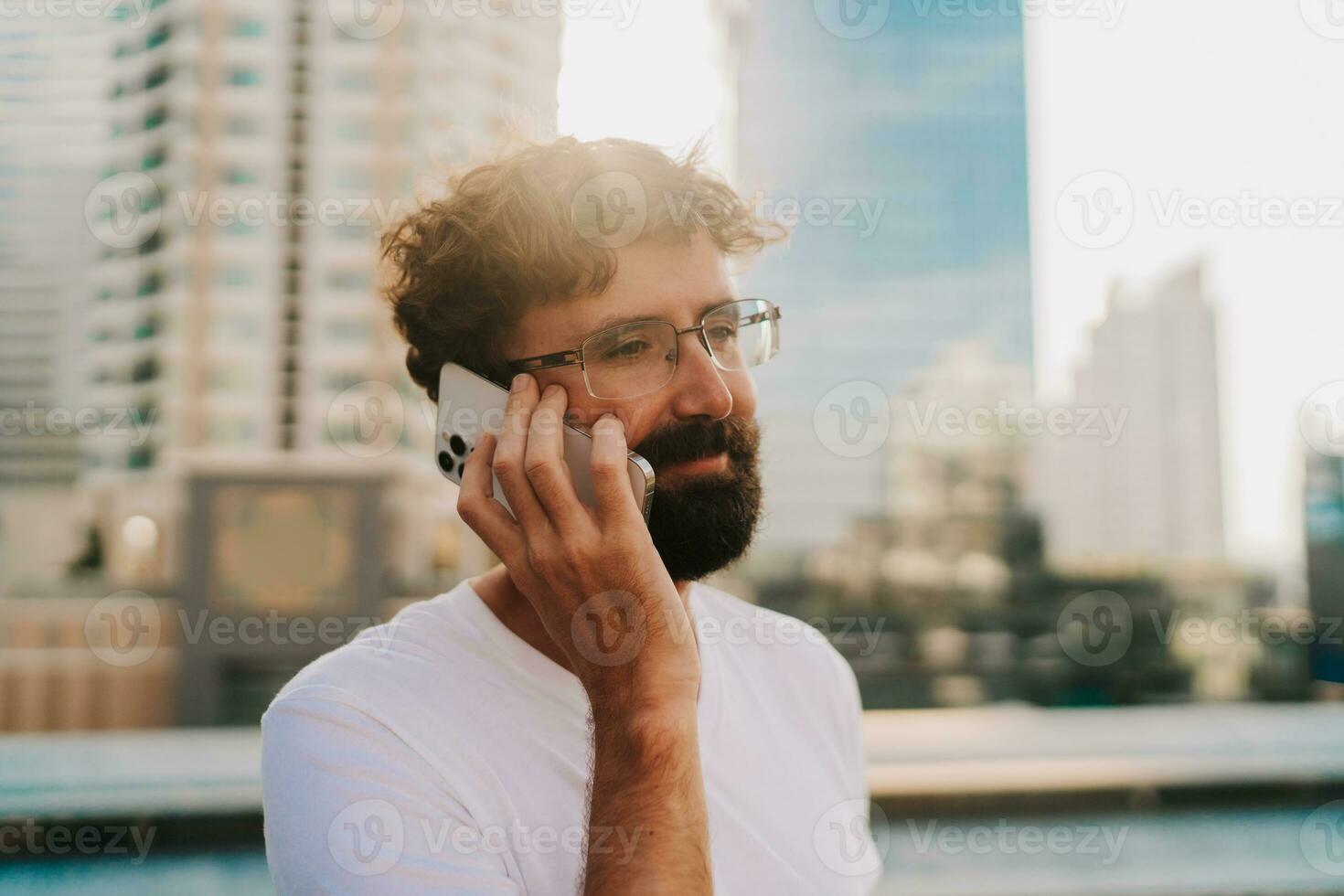 stilig man med skägg talande förbi mobil telefon medan gående i stor modern stad med skyskrapor. utomhus på solnedgång . bär vit t skjorta. foto