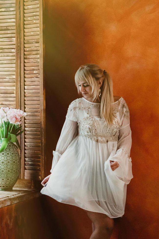 ung blond kvinna i vit klänning som dansar vid fönstret, färska rosa blommor bukett i vacker vas, ljus sienna vägg i bakgrunden. romantisk, kärlek, lycka koncept foto
